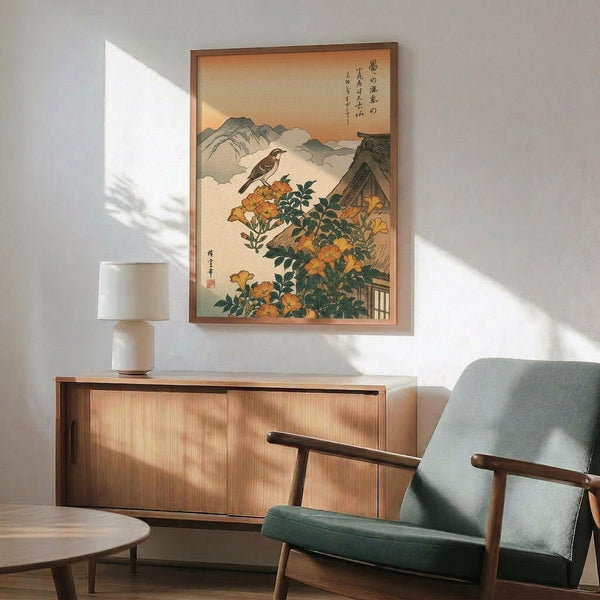 May include: Japanese woodblock print artwork displayed in modern living room with mid-century furniture. The framed poster shows a brown bird perched on orange trumpet vine flowers with mountains and traditional architecture in background. Warm peachy sky with Japanese calligraphy. Wooden sideboard below holds white ceramic lamp, sage green upholstered chair with wooden arms nearby, natural sunlight casting shadows on white wall.
