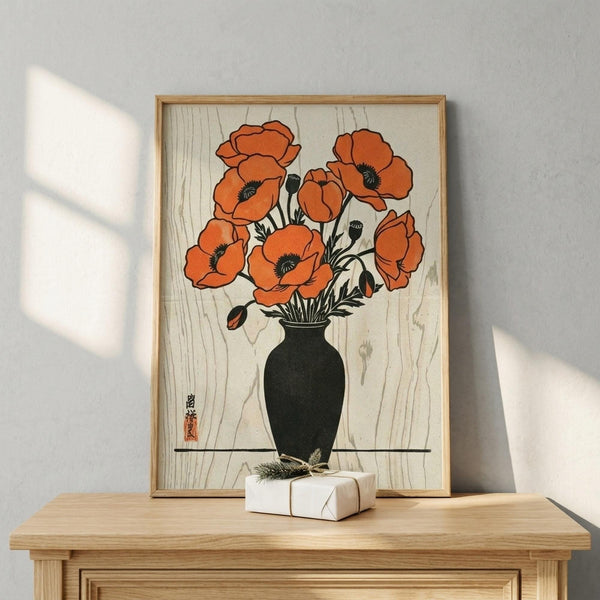 May include: Framed Japanese woodblock-style print showing orange poppies in a dark green vase, displayed on a light wood surface against a white wall with window shadows. The artwork features bold orange blooms with black centers on a cream background with wood grain texture. A small wrapped gift sits beside the thin wooden frame.