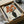 A Japanese woodblock print artwork showing orange poppies in a dark vase, displayed in a black frame inside an open cardboard shipping box with protective tissue paper. The print features bold orange blooms with black centers against a light wood-grain background, created in traditional ukiyo-e style with clean lines and flat colors.