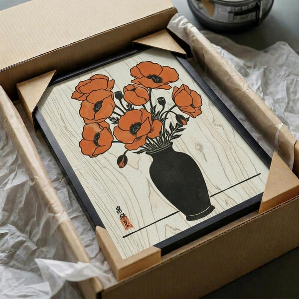 A Japanese woodblock print artwork showing orange poppies in a dark vase, displayed in a black frame inside an open cardboard shipping box with protective tissue paper. The print features bold orange blooms with black centers against a light wood-grain background, created in traditional ukiyo-e style with clean lines and flat colors.