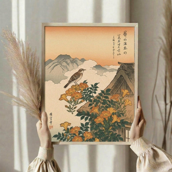 May include: Person in cream ruffled sleeves holding framed Japanese woodblock print showing orange trumpet vine flowers, brown bird perched on flowering branch, traditional thatched roof building, mountains and clouds in background with peachy sky. Dried pampas grass visible to left. Print features Japanese calligraphy and artist's red seal stamp in traditional ukiyo-e style with muted earth tones.