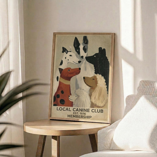 May include: Framed vintage-style poster featuring illustrated dogs of various breeds including dalmatian, bull terrier, and poodle on cream background, displayed on round wooden side table next to beige upholstered chair with plant leaves visible in soft natural lighting