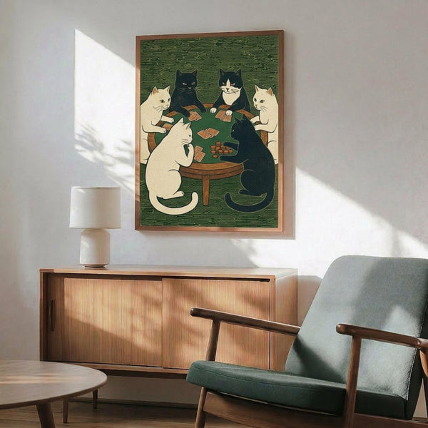 May include: Framed poster print in wooden frame hanging on white wall above mid-century modern wooden credenza with table lamp. Green fabric armchair and round coffee table visible in foreground. Artwork shows five cats in cream, black, and tuxedo colors sitting around green felt card table playing poker, rendered in woodblock print style with textured green background.