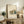 May include: Framed Bauhaus poster featuring minimalist architectural illustration of modernist building with clean geometric lines in muted green and orange tones, displayed on rustic wooden console table alongside potted fern, ceramic vase, and books in warm-toned living room setting with cream upholstered chair.