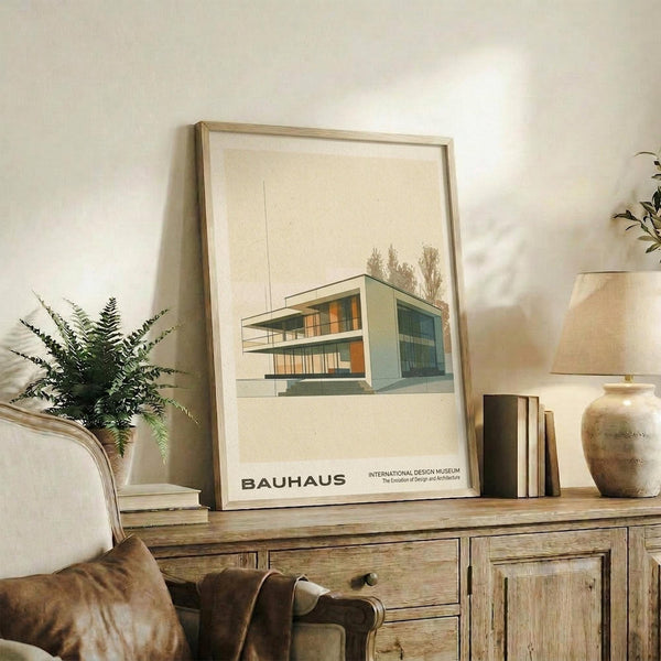 May include: Framed Bauhaus poster featuring minimalist architectural illustration of modernist building with clean geometric lines in muted green and orange tones, displayed on rustic wooden console table alongside potted fern, ceramic vase, and books in warm-toned living room setting with cream upholstered chair.