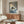May include: Framed artwork showing a black cat with green eyes reading an open book while sitting on striped bedding in blue and white tones. The painting is displayed above a wooden sideboard with sliding doors in a modern living room with cream walls, white upholstered chair, glass-top coffee table, and minimalist lighting fixtures.
