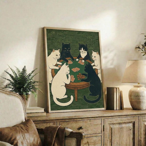 May include: Framed artwork showing five cats sitting around a round wooden table playing cards in woodblock print style. White and black cats with detailed expressions hold cards while poker chips sit on green table surface. Green textured background. Light wood frame displayed on rustic wooden sideboard with books, ceramic vase, and fern plant in bright, neutral-toned living room setting.