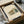 May include: Framed Bauhaus-style architectural print featuring a modernist building design in muted green, orange and beige tones, displayed in a black frame within an open cardboard shipping box surrounded by white tissue paper and brown paper tubes on a neutral surface.