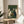 May include: Framed vintage-style poster featuring five cats sitting around a wooden table playing cards on green background. The illustration shows white, black, and tuxedo cats in woodblock print style with visible wood grain texture. The poster sits on a round wooden side table next to a beige upholstered chair with white throw pillow, with plant leaves and window light visible in the background.