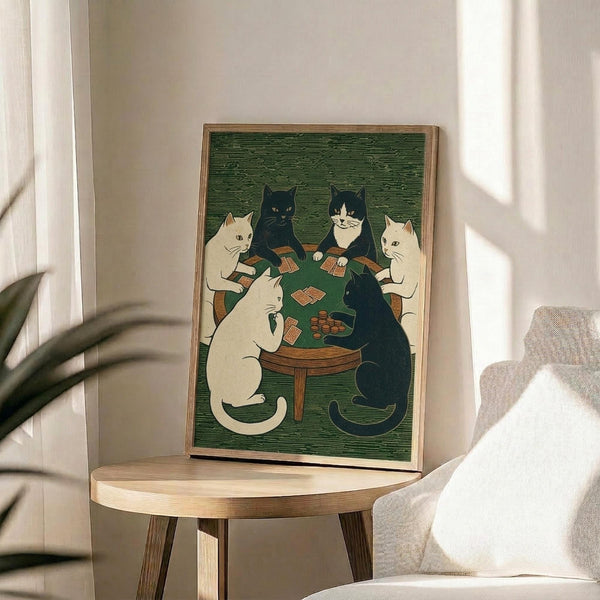 May include: Framed vintage-style poster featuring five cats sitting around a wooden table playing cards on green background. The illustration shows white, black, and tuxedo cats in woodblock print style with visible wood grain texture. The poster sits on a round wooden side table next to a beige upholstered chair with white throw pillow, with plant leaves and window light visible in the background.