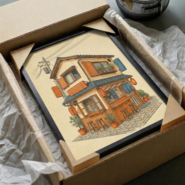 May include: Framed artwork depicting a cozy two-story Japanese ramen shop with warm wood exterior, orange signage with Japanese characters, potted plants, and stone pathway. The illustration shows detailed architectural features including a tiled roof, power lines, and traditional storefront elements. The print has a cream background and is displayed in a black frame, partially unpacked from brown cardboard packaging with white tissue paper visible around the edges.