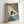May include: Framed artwork leaning against white wall showing black cat reading small book while sitting in bed with blue striped pillows and blankets. Cozy bedroom scene with warm lighting from lamp, bookshelves visible in background. Light wood frame contains illustration in muted blue and cream tones with realistic, detailed art style.