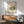 May include: Framed Bauhaus architectural poster showing modernist building in muted green and orange tones, displayed above wooden sideboard with white ceramic lamp. Mid-century armchair with sage green upholstery and walnut arms sits nearby. Natural sunlight creates geometric shadows on white wall, complementing the clean lines and warm wood furniture in this minimalist interior setting.
