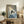 May include: Framed artwork showing a black cat reading a book while sitting in a striped blue and white bed, displayed on a rustic wooden sideboard alongside decorative pottery, books, and a fern plant in a cozy living room setting with warm lighting and neutral walls.