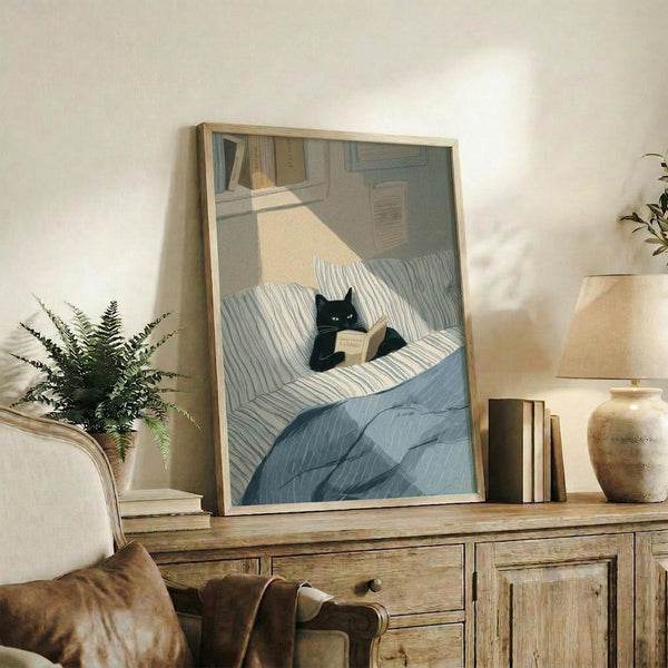May include: Framed artwork showing a black cat reading a book while sitting in a striped blue and white bed, displayed on a rustic wooden sideboard alongside decorative pottery, books, and a fern plant in a cozy living room setting with warm lighting and neutral walls.