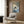 May include: Framed artwork showing a black cat reading a book while lying on striped bedding in blue and white tones, displayed on wood-paneled wall above a beige upholstered chair with geometric pillow. Modern floor lamp and side table with open book visible in contemporary neutral-toned room setting with curtained window.