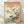 May include: Japanese woodblock print artwork in light wood frame leaning against white wall on wooden floor. Features pink cherry blossoms on brown branch with blue bird perched among flowers, Mount Fuji with snow-capped peak and clouds in background, flowing water at bottom with green leaves. Warm beige and orange sky with vertical Japanese text calligraphy on right side.