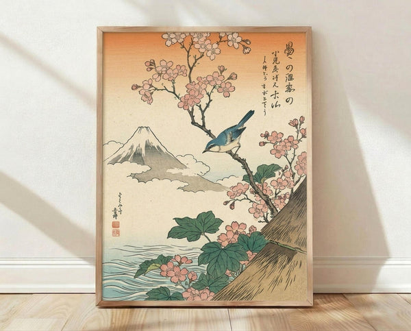 May include: Japanese woodblock print artwork in light wood frame leaning against white wall on wooden floor. Features pink cherry blossoms on brown branch with blue bird perched among flowers, Mount Fuji with snow-capped peak and clouds in background, flowing water at bottom with green leaves. Warm beige and orange sky with vertical Japanese text calligraphy on right side.