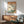 May include: Japanese woodblock print artwork in wooden frame showing Mount Fuji, cherry blossoms, and blue bird, displayed above mid-century modern walnut credenza with ceramic table lamp, next to sage green upholstered armchair in bright sunlit room with white walls.