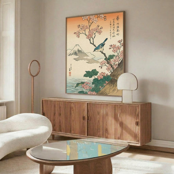 May include: Japanese woodblock print artwork hanging above wooden sideboard in modern living room. Print features Mount Fuji, pink cherry blossoms, blue bird, and Japanese text on cream background in thin frame. White curved chair and glass-top coffee table with walnut base visible. Warm natural lighting creates shadows on beige walls.