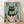 May include: Framed poster print leaning against white wall on wooden floor. Artwork shows black cat with yellow eyes sitting on pink toilet, reading green book. Turquoise tiled bathroom background with terra cotta plant pots containing cacti and succulents on shelves and floor. Illustration has textured, hand-drawn style with speckled details. Natural wood frame surrounds the colorful print in this cozy room mockup scene.