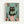 Whimsical illustration of a black cat with yellow eyes sitting on a pink toilet while reading a green book. The bathroom scene features turquoise tile walls, potted plants on shelves and floor including a cactus and leafy plants in terracotta and pink pots. The artwork has a folk art style with textured brushstrokes and is displayed in a light wood frame against a white wall.