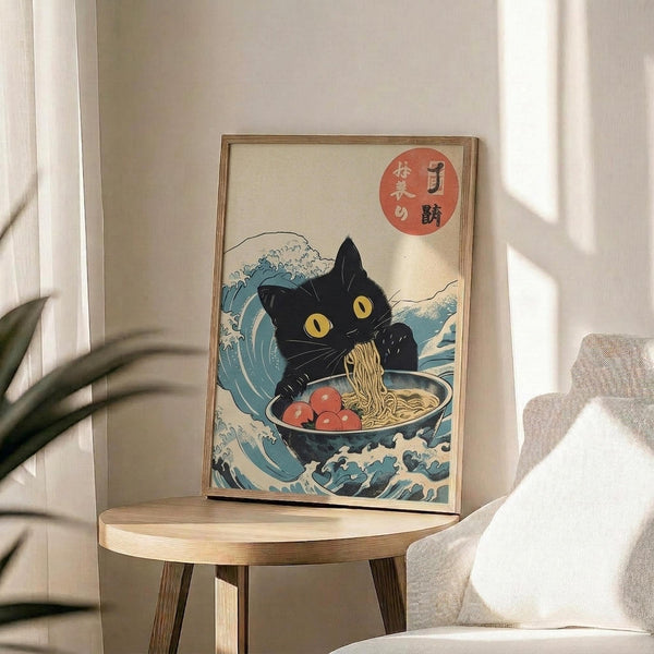 May include: Framed poster print displayed on wooden side table next to beige chair and plant. Artwork shows black cat with yellow eyes eating ramen from bowl, surrounded by blue ocean waves in Japanese ukiyo-e style. Red circular seal with Japanese characters in upper right corner. Light wood frame, cream colored background, natural lighting creates soft shadows.