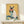 May include: Framed artwork showing a Siamese cat wearing pink sunglasses and light blue shirt, holding a watermelon slice. The cat has cream and dark brown coloring against a warm yellow background with teal accents. Orange slices sit at the bottom. The piece is displayed on a light wood surface next to a small white wrapped gift, leaning against a cream wall.