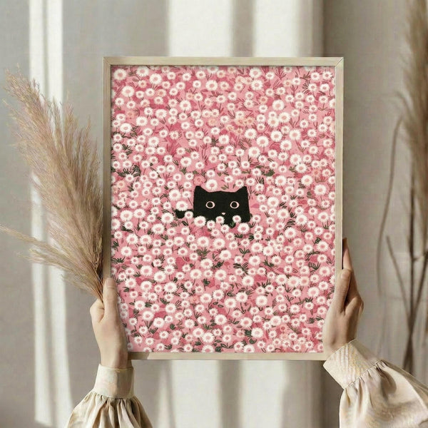 May include: Person holding framed artwork showing black cat silhouette peeking through dense field of pink and white daisy flowers. Thin light wood frame, held against neutral curtained window background. Dried wheat stems visible on left side. Cozy home interior setting with soft natural lighting.