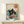 May include: Framed poster print displayed on light wood furniture against white wall. Artwork shows black cat with yellow eyes reaching into ramen bowl with red ingredients, surrounded by blue ocean waves in Japanese ukiyo-e style. Red circular text element in upper right corner. Natural wood frame, cream matting. Small white wrapped gift box with greenery sits in front of frame.