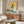 May include: Framed poster artwork displayed above a wooden sideboard in a modern living room setting. The artwork shows a white and dark Siamese cat wearing round sunglasses, holding a watermelon slice, painted in vibrant colors against a yellow background. The room features a white upholstered chair, glass coffee table, small plant, and mirror on the sideboard.