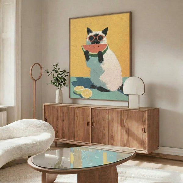 May include: Framed poster artwork displayed above a wooden sideboard in a modern living room setting. The artwork shows a white and dark Siamese cat wearing round sunglasses, holding a watermelon slice, painted in vibrant colors against a yellow background. The room features a white upholstered chair, glass coffee table, small plant, and mirror on the sideboard.