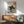 May include: Japanese-style artwork print showing black cat with yellow eyes eating ramen noodles from bowl with red garnish, surrounded by blue ocean waves in ukiyo-e style. Framed poster hangs above mid-century modern wood credenza with white lamp, next to sage green upholstered armchair in bright, minimalist living room setting.