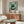 May include: Whimsical illustration of black cat with yellow eyes reading green book while sitting on pink toilet, surrounded by potted plants on mint green tiled bathroom wall. Artwork displayed above mid-century modern walnut credenza in living room with white curved chair, brass floor lamp, and oval glass coffee table on light wood floors.