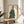 May include: Framed art print displayed on wooden side table next to beige upholstered chair. The artwork features a stylized figure with leopard-spotted face wearing sunglasses and a flowing polka-dotted garment in sage green with black dots. Natural lighting creates soft shadows across the neutral-toned room setting with plant leaves visible in corner.