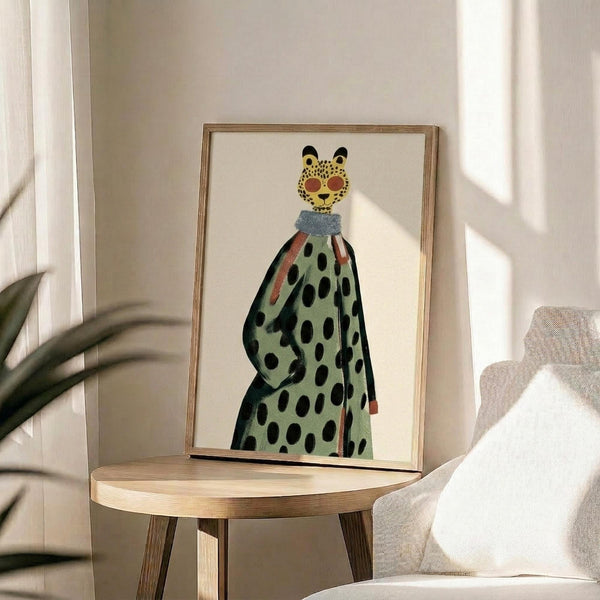 May include: Framed art print displayed on wooden side table next to beige upholstered chair. The artwork features a stylized figure with leopard-spotted face wearing sunglasses and a flowing polka-dotted garment in sage green with black dots. Natural lighting creates soft shadows across the neutral-toned room setting with plant leaves visible in corner.
