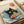 Japanese-style woodblock print artwork featuring a black cat with yellow eyes sitting in a ramen bowl surrounded by blue ocean waves reminiscent of Hokusai's Great Wave. The cat holds chopsticks while noodles and red garnishes float in the bowl. A red circular seal with Japanese characters appears in the upper left corner. The print is displayed on cream-colored paper in a wooden frame against white surfaces.