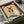 May include: Framed poster print being unpacked from cardboard shipping box with protective tissue paper. Artwork shows white and black Siamese cat wearing red sunglasses, eating watermelon slice against yellow and turquoise background with orange slices below. Black frame sits in brown cardboard box on white surface next to tape dispenser.