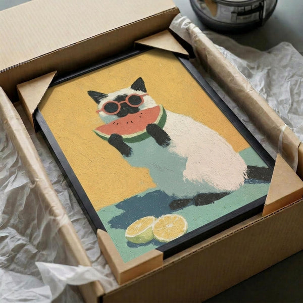 May include: Framed poster print being unpacked from cardboard shipping box with protective tissue paper. Artwork shows white and black Siamese cat wearing red sunglasses, eating watermelon slice against yellow and turquoise background with orange slices below. Black frame sits in brown cardboard box on white surface next to tape dispenser.