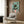 May include: Whimsical illustration of black cat with yellow eyes reading green book while sitting on pink toilet, surrounded by potted plants on turquoise tiled bathroom wall. Framed artwork displayed above beige upholstered armchair with geometric pillow in modern living room with wood paneling, brass floor lamp, and dark side table.