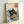 Framed poster print featuring a black cat with yellow eyes sitting in a ramen bowl filled with noodles and red garnishes, surrounded by stylized blue ocean waves in traditional Japanese ukiyo-e art style. The artwork has a cream background with Japanese characters in a red circular seal. The print is displayed in a light wood frame leaning against a white wall.
