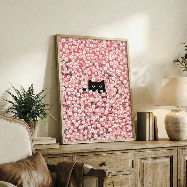 May include: Framed poster with pink floral pattern and small black cat peeking through displayed on rustic wooden sideboard in cozy living room setting with fern plant, books, ceramic vase, and beige upholstered furniture visible in warm natural lighting