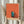 May include: Hands holding framed artwork featuring orange geometric walls with small doorway containing silhouette of person reading, partial moon visible, minimalist contemporary style. Light wood frame against neutral curtained background with dried pampas grass visible on left side.