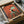 May include: Framed poster print in black frame with cardboard packaging, showing vintage-style artwork of black and white tuxedo cat sitting on turquoise wooden chair surrounded by small dark birds against warm orange-red textured background. Frame sits in shipping box with protective tissue paper on table surface.