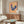 May include: Colorful folk art poster featuring a blue hen with red comb and pink flowers on its body, displayed against yellow and pink striped background. Shown framed on wall above walnut credenza in modern living room with white curved chair, glass coffee table, and table lamp.