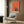 May include: Orange and beige abstract poster with small blue window scene featuring a silhouette reading, displayed in light wood frame on wood-paneled wall above cream upholstered armchair with geometric pillow, brass floor lamp and dark side table with open book nearby