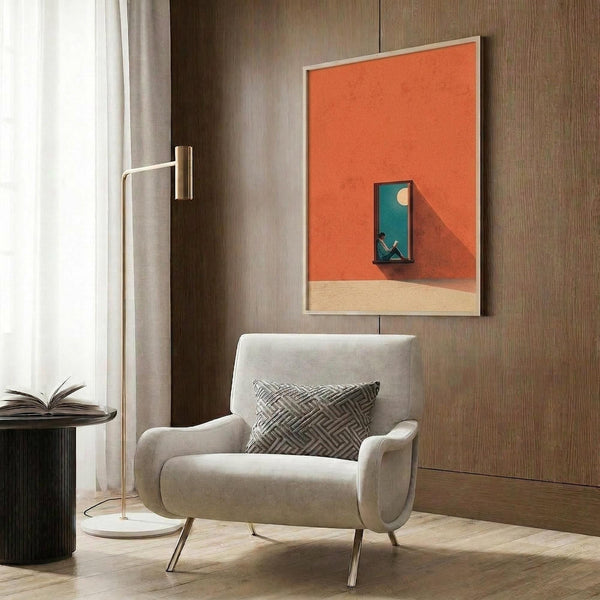 May include: Orange and beige abstract poster with small blue window scene featuring a silhouette reading, displayed in light wood frame on wood-paneled wall above cream upholstered armchair with geometric pillow, brass floor lamp and dark side table with open book nearby
