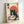 Framed poster in light wood frame leaning against white wall, featuring a black cat with white eyes eating ramen noodles with chopsticks. The cat sits behind a decorative bowl with an egg and noodles, alongside a small bottle. A large orange circle resembling the sun fills the background. Japanese text appears in the upper left and right corners. The artwork uses a vintage Japanese woodblock print style with cream, orange, and black colors on textured paper background.
