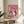 May include: Whimsical artwork in gold frame showing two white geese wearing sunglasses and colorful scarves sharing pizza and red wine on pink background. Frame sits on round wooden side table next to beige upholstered chair with white throw pillow. Soft natural lighting creates shadows on neutral wall. Green plant leaves visible in corner.