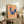 May include: Framed folk art poster featuring a blue hen with red comb and pink floral pattern on body, set against yellow and pink striped background. Artwork displayed on rustic wooden sideboard next to fern plant, books, and ceramic vases in neutral-toned living room setting.